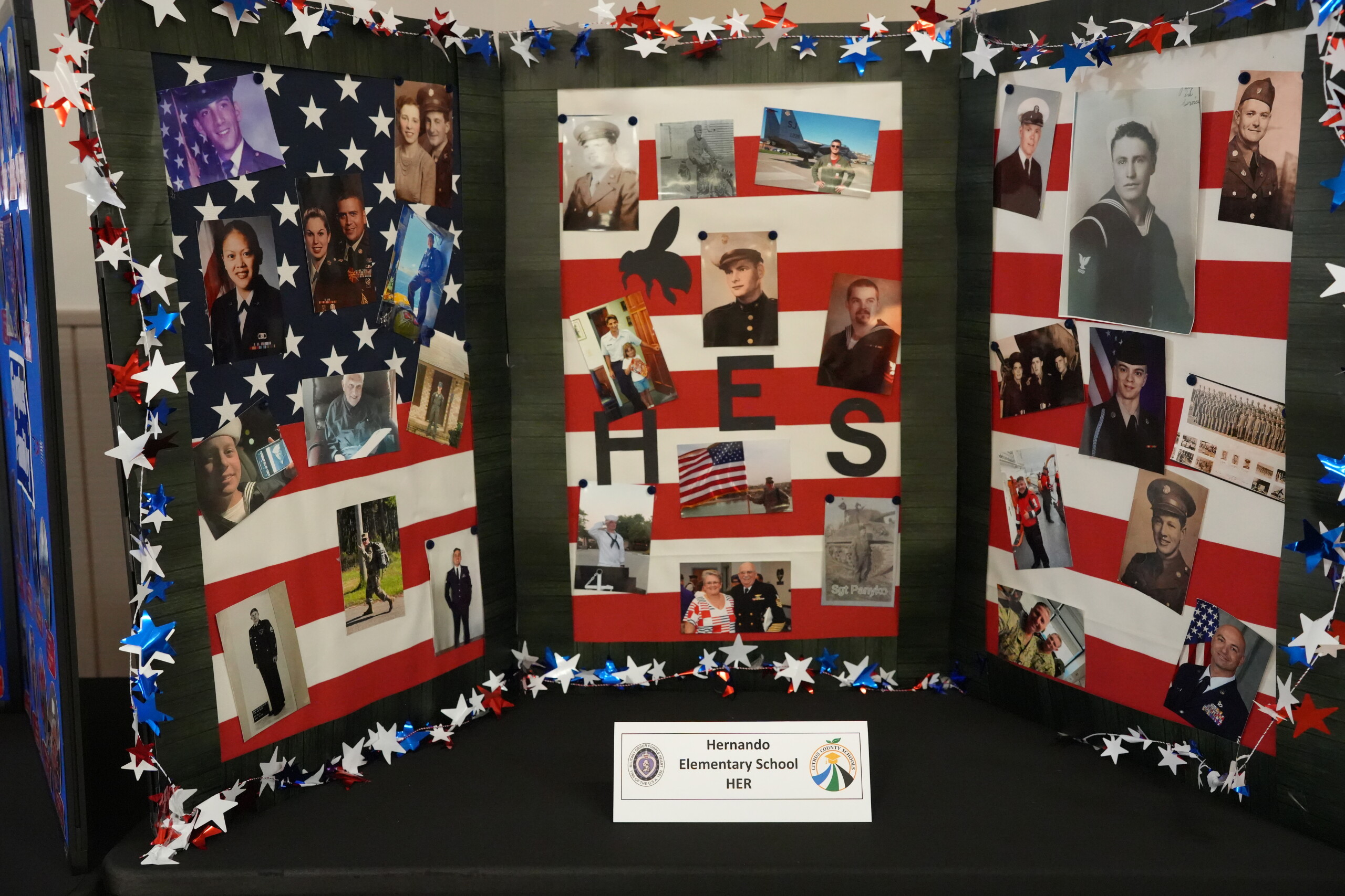 A tri-fold display board decorated with American flags and red, white, and blue star garlands features many photographs of service members.